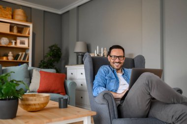 Genç girişimci gülümsüyor ve evde otururken bilgisayarda çalışıyor. Erkek serbest çalışan, merkez ofisteki kablosuz bilgisayar üzerinde çalışırken kameraya mutlu bir şekilde bakıyor.