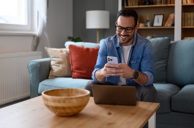 Sofrada otururken akıllı telefondan mesaj atan dizüstü bilgisayarlı gülümseyen genç işadamı. Ev ofisinden çalışırken kablosuz bilgisayar ve cep telefonuyla telekomünikasyon yapan erkek girişimci.