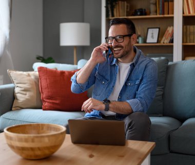Neşeli erkek serbest çalışan cep telefonuyla konuşuyor ve kanepede otururken dizüstü bilgisayarla çalışıyor. Genç işadamı ev ofisinden çalışırken cep telefonuyla iletişim kuruyor.
