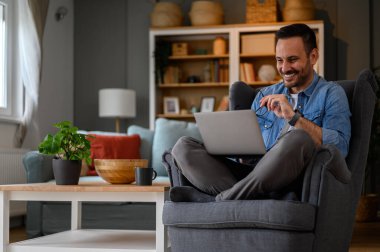 Dizüstü bilgisayarda çalışan mutlu erkek bilgisayar programcısı koltukta bacak bacak bacak üstüne atarak rahatça oturuyor. Ofiste uzaktan çalışırken kablosuz bilgisayar kullanan genç, gülümseyen bir serbest yazar.