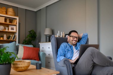 Koltuğunda rahat bir şekilde oturan ve dizüstü bilgisayarla çalışan neşeli genç bir iş adamı. Ofiste dinlenirken kablosuz bilgisayardan içerik kodlayan serbest çalışan erkek.