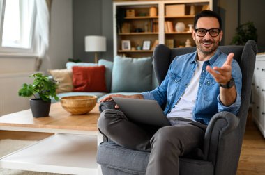 Ev ofisindeki koltukta otururken kameraya el kol hareketi yapan mutlu bir iş adamı.