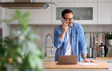 Evde mutfak tezgahında dizüstü bilgisayar kullanırken akıllı telefondan konuşan kendinden emin bir erkek profesyonel.