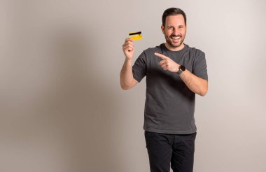 Kredi kartını arka planda gösteren ve işaret eden alışverişçi bir gencin portresi. Yakışıklı erkek müşteri gülüyor ve online alışveriş ve elektronik bankacılık öneriyor.