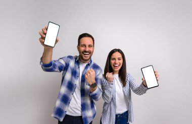 Heyecanlı kız arkadaşın portresi, erkek arkadaşının gülüşü ve cep telefonlarını gösterirken yumruklaşması. Mobil uygulamaları destekleyen ve arka plandaki başarıyı kutlayan kendinden geçmiş genç çift.