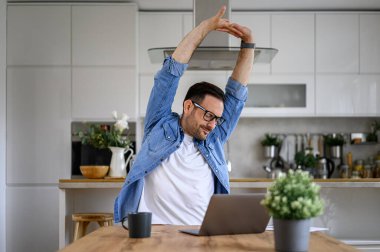 Yorgun erkek girişimci ofis masasındaki laptopta fazla mesai yaparken kollarını uzatıyor.
