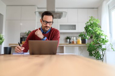 Ciddi bir iş adamı laptopta çalışıyor ve not defterine notlar yazıyor.