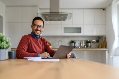 Ev ofisinden çalışan, bilgisayarlı ve not defterli neşeli yakışıklı adam kameraya gülümsüyor.