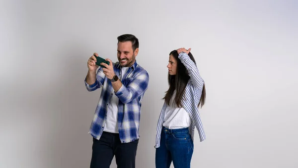背景にスマホで興奮した彼氏がビデオゲームをしているのを猛烈に見つめている手に頭を持つ彼女 携帯電話を使用して陽気な男を見て深刻な女性 — ストック写真