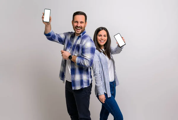 Joyful young man and woman showing blank screen of smart phones and promoting e-commerce apps. Cheerful couple dressed in casuals advertising over cellphones on background