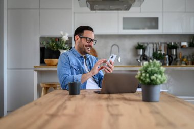 Sosyal medyayı akıllı telefondan kullanan neşeli, yakışıklı iş adamı ofis masasında dizüstü bilgisayarla.