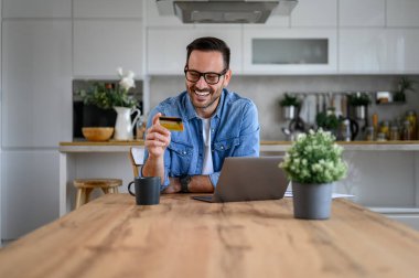 Genç işadamının elinde kredi kartı var ve evdeki ofiste bilgisayardan alışveriş yapıyor.