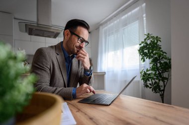 Takım elbiseli odaklanmış iş adamı çeneye dokunuyor ve ofisteki masada laptopun üzerinde çalışıyor.