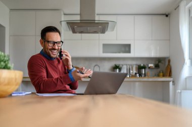 Zeki telefondan konuşan ve ofisteki masadaki laptopa bakan neşeli serbest çalışan bir erkek.