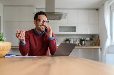 Neşeli genç girişimci telefonda konuşuyor ve ofis masasında dizüstü bilgisayar kullanıyor.
