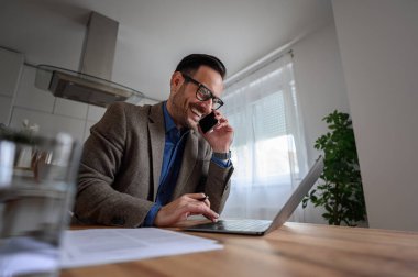 Mutlu iş adamının akıllı telefonla konuşması ve ev ofisinin bilgisayarında çalışmasının düşük açılı görüntüsü.