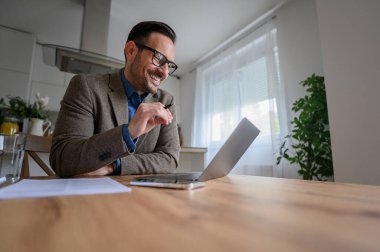 Takım elbise giymiş şık bir iş adamı. Evde bilgisayarla görüşmeler yapıyor ve gülümsüyor.