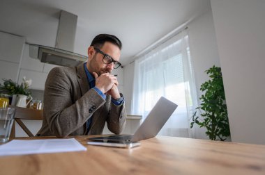 Odaklanmış erkek profesyonel laptopa bakıyor ve evden çalışırken iş stratejileri düşünüyor.