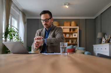 Ciddi bir iş adamının portresi. Ev bilgisayarında çalışırken kol saatini kontrol ediyor.