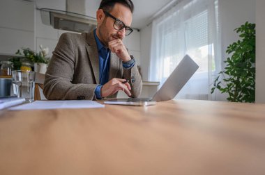 Takım elbise giymiş, çeneye dokunan ve ofisteki masada dizüstü bilgisayar kullanan odaklanmış genç bir iş adamı.