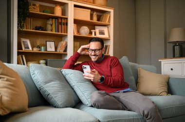 Mutlu genç işadamı oturma odasındaki koltukta akıllı telefondan sosyal medya pazarlaması yapıyor.