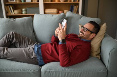 Genç işadamı oturma odasındaki kanepede dinlenirken akıllı telefondan gülümsüyor ve mesajlaşıyor.