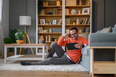 Gülümseyen yakışıklı girişimci evdeki ofiste halının üzerinde otururken dijital tablet üzerinde çalışıyor.