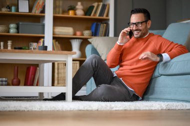 Genç işadamı gözlük takıyor ve oturma odasında dinlenirken akıllı telefondan konuşuyor.