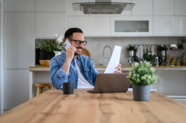 Akıllı telefondan konuşan ve ofis masasında dizüstü bilgisayarla doküman okuyan mutlu iş adamı.