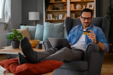 Bilgisayarlı ve kredi kartlı yakışıklı iş adamı online alışveriş yapıyor ve evdeki koltukta oturuyor.