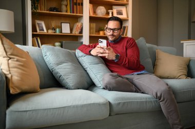 Yakışıklı genç adam oturma odasındaki kanepede dinlenirken akıllı telefonuyla sosyal medyada geziniyor.