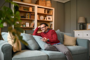 Gülümseyen girişimci ev ofisinin oturma odasında koltukta dinlenirken cep telefonu kullanıyor.