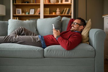 Mutlu erkek girişimci oturma odasındaki kanepede yatarken akıllı telefondan mesajları kontrol ediyor.