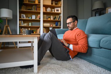 Gözlüklü, işine odaklanmış, dizüstü bilgisayarda araştırma yapan, halı üzerinde kanepede dinlenen bir erkek girişimci.