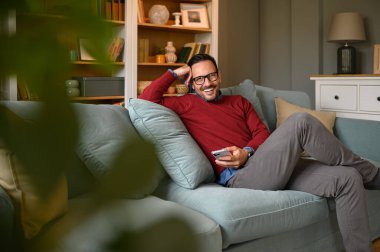 Yakışıklı genç girişimci oturma odasındaki kanepede dinlenirken gülümseyen ve akıllı telefonu kaydıran.