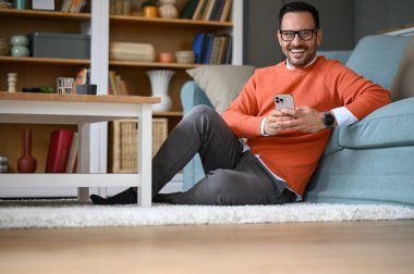 Genç işadamı kameraya gülümsüyor ve oturma odasında halıda otururken akıllı telefon kullanıyor.