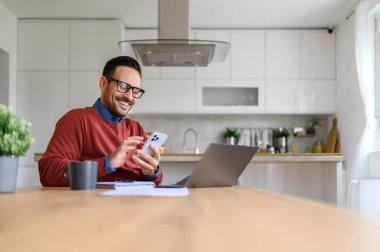 Dahili ofiste bilgisayarla çalışırken akıllı telefondan mesaj atan yakışıklı bir iş adamı.