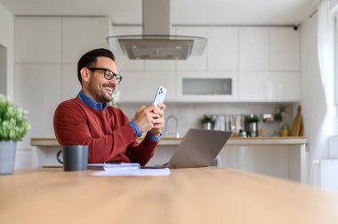 Mutlu yakışıklı iş adamı evdeki masada dizüstü bilgisayar kullanırken internetten akıllı telefon mesajları atıyor.
