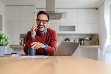 Dahili ofiste dizüstü bilgisayarla otururken akıllı telefondan konuşan neşeli iş adamı.