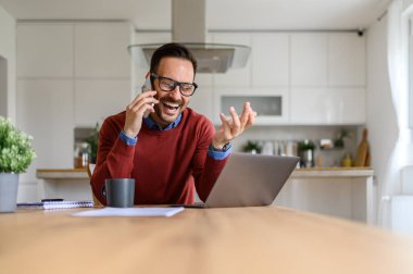 Neşeli erkek serbest çalışan akıllı telefondan iş görüşmesi yapan evden laptopla çalışırken.