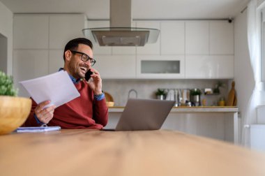 Hesap çizelgesi olan erkek girişimci evde çalışırken akıllı telefondan iş görüşmesi yapıyor.