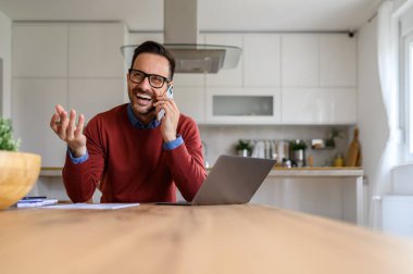 İş adamı ofisteki masasında otururken cep telefonuyla konuşuyor ve neşeyle gülüyor.