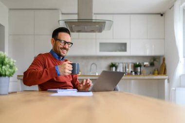 Gülümseyen işadamı, ofisteki masada bilgisayardan yapılan video görüşmesi sırasında kahve içiyor.