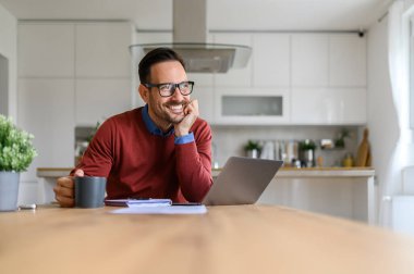Düşünceli, gülümseyen bir işadamı, elleri çenesinde, ofis masasında otururken gözlerini kaçırıyor.