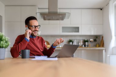 Ofisteki masada dizüstü bilgisayar kullanırken akıllı telefondan neşeli bir şekilde konuşan bir erkek.