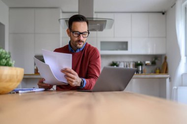 Finansal raporları elinde tutan ve evde çalışırken bilgisayarını kullanan ciddi bir mali danışman.