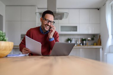 Evrakları elinde tutan ve evde çalışırken akıllı telefondan konuşan mutlu genç işadamı.