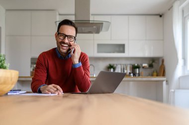 Neşeli, genç, serbest çalışan bir erkek. Masa başında çalışırken telefonla konuşuyor.