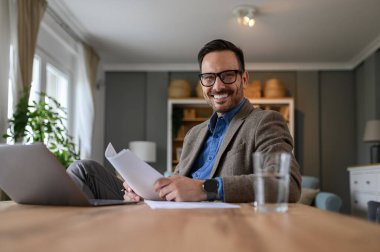 Elinde belgelerle gülümseyen zarif bir işadamı ve evde çalışırken kameraya bakıyor.