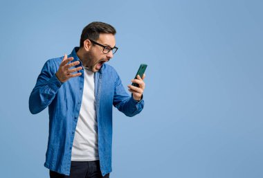 Erkek profesyonel, mavi arka planda akıllı telefondan mesaj okurken öfkeyle bağırıyor.